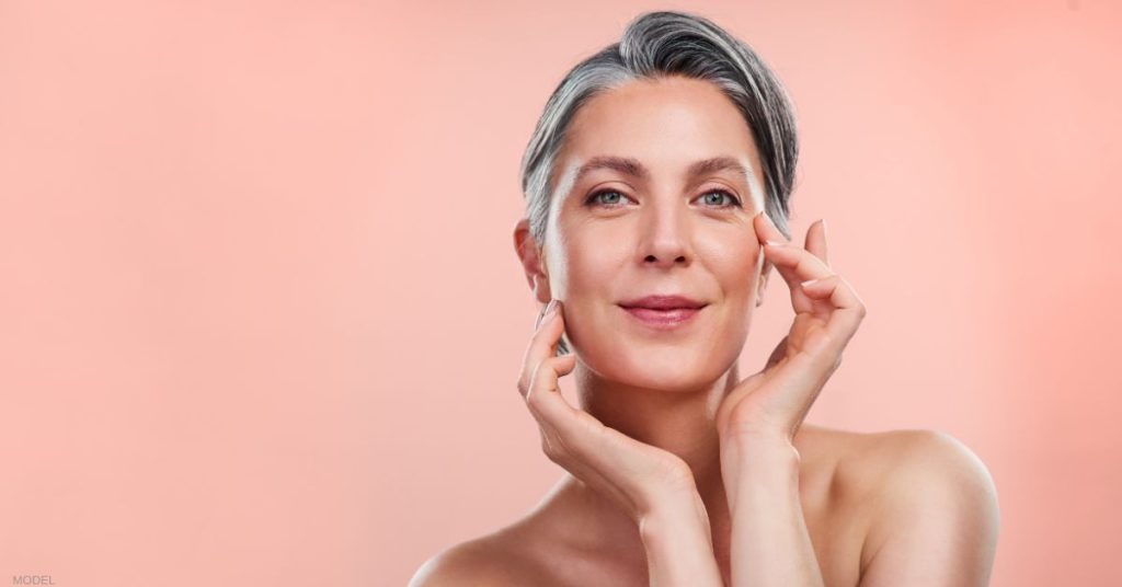 Woman with gray hair posing for the camera, gently touching the area around her eye (MODEL)