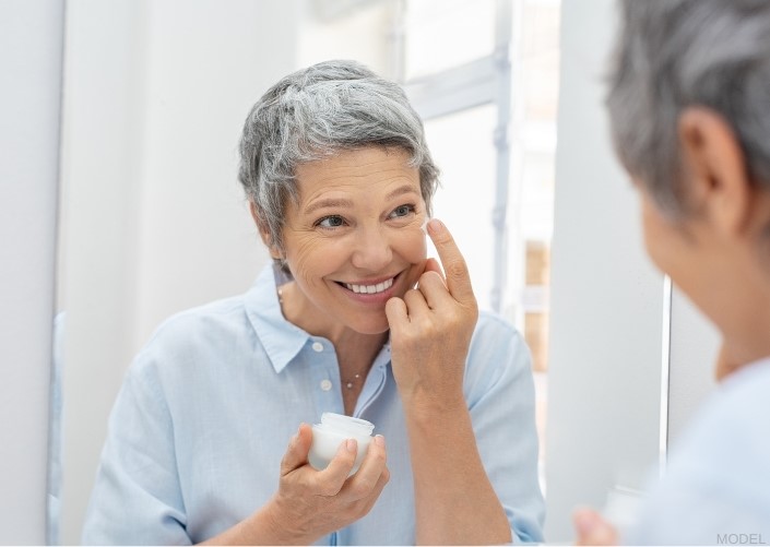 Mature woman looking in the mirror while applying cream to their face (MODEL)